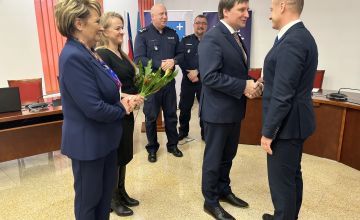 Zdjęcie przedstawiające sześć osób. Dwie kobiety stojące z przodu jedna z nich trzymie kwiaty. Przed nimi jeden meższczyzna ściska rękę drugiemu w geście gratulacyjnym. W tle stoi dwóch policjantów w mundurach. Zdjęcie wykonane na sali konferencyjnej na scianie widać element wiszącego godła.